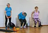 Tre eldre kvinner trenar styrke på kasser i ein gymsal med utstyr på golvet.