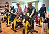 Eldre kvinner trenar saman på gule spinningsyklar i ein sal med lyst tre­golv.