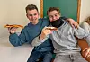 To ungdomar sit tett saman ved eit bord og held opp pizzastykke mot kameraet.