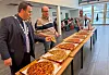 Fleire vaksne deler ut fleire store pizzaer på bord i ein skulekantine.