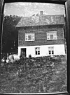 Historisk svart-kvitt foto av eit nyoppført trehus med hage og to personar framfor.