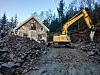 Beltegravemaskin står på ein steinrik gardsplass framfor eit hus i skogkledd terreng.