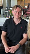 Ung mann i svart poloskjorte sit i eit kafé- eller restaurantmiljø.