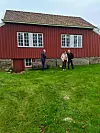 Tre personar står framfor ein raudmåla trebygning med kvite vindauge og grønt gras.