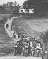 Gruppe motorsyklistar på ein grusveg ved eit hus og skogkledd landskap.