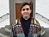 Uidentifisert prest i fargerik cardigan står framfor ein kyrkjeinngang.