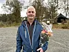 Smilande mann i blå jakke held ein blomsterbukett på ein grusplass.