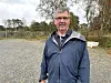 Mann i blå jakke står på ein grusplass med skog og tre i bakgrunnen.