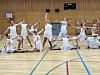 Gruppe utøvarar i kvite klede utfører ein synkronisert oppvisning i ein gymsal.